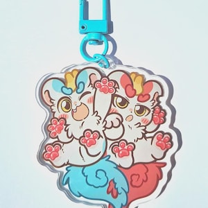 Może przedstawiać: Przezroczysty akrylowy brelok do kluczy z dwoma kreskówkowymi stworzeniami przypominającymi koty z białym futrem, różowymi poduszkami łap i niebieskimi i czerwonymi ogonami. Brelok ma jasnoniebieskie zapięcie.