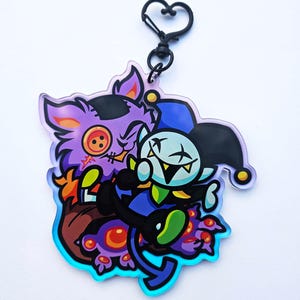 Könnte beinhalten: Ein farbenfroher Acryl-Anhänger mit zwei Zeichentrickfiguren. Einer ist ein lila katzenartiges Wesen mit Knopfauge und gestickten Details. Der andere ist eine Narrenfigur in Blau und Schwarz. Der Anhänger hat einen herzförmigen Verschluss.
