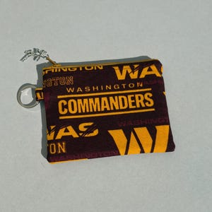 May include: A small, rectangular coin purse with a zipper closure. The purse is patterned with the words "WASHINGTON COMMANDERS" in gold and white on a maroon background. It has a silver key ring attached.