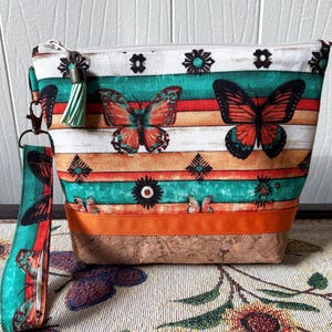 May include: A rectangular cosmetic bag with a butterfly print in orange, teal, and white stripes. The bag has a cork base, a zipper closure, and a matching wrist strap. The bag is decorated with a green and white tassel.