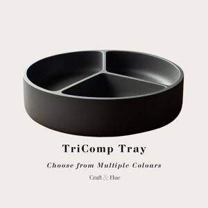 Può includere: Un vassoio circolare nero opaco con tre scomparti interni distinti separati da divisori a forma di Y. Il testo sotto il vassoio recita TriComp Tray, Choose from Multiple Colours e Craft & Hue in un carattere serif scuro e pulito.