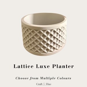 Peut inclure: Un pot de fleurs cylindrique blanc cassé avec un motif en losanges en relief sur l'extérieur. Le texte ci-dessous indique Lattice Luxe Planter, Choose from Multiple Colours et Craft & Hue. Le pot présente une ouverture circulaire large.