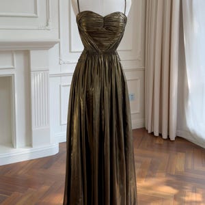 Könnte beinhalten: Ein bodenlanges, metallisch bronzefarbenes Kleid mit herzförmigem Ausschnitt, dünnen Spaghettiträgern und einem gerafften Oberteil. Der Rock ist an der Taille gerafft und erzeugt eine volle, fließende Silhouette mit vertikalen Falten.