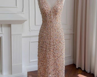 Puede incluir: Un vestido de noche sin mangas y largo hasta el suelo sobre un maniquí. El vestido es de color melocotón claro y está cubierto de lentejuelas. Tiene un escote en V y una silueta ajustada. El vestido se exhibe frente a una pared blanca y una ventana con cortinas transparentes.