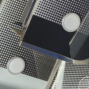 May include: Close-up of a silver and black accessory with a geometric design. The accessory features a black rectangular center, silver trim, and two white circular accents. The background is a pattern of white dots on a black surface.