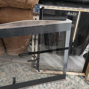 May include: A close-up of a black and silver glass fireplace door with a silver frame. The door is partially open, revealing the interior of the fireplace. The door is made of tempered glass and has a safety latch.