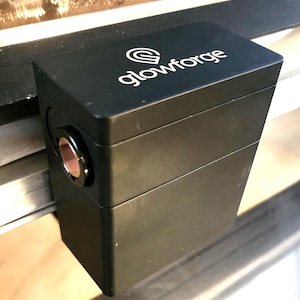 May include: A black rectangular device with the word "glowforge" in white script. The device has a circular opening with a copper-colored interior. The device is mounted on a metal rail.