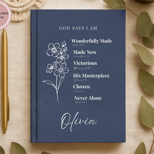 Puede incluir: Un diario de tapa dura azul marino con una ilustración floral de líneas blancas y el texto GOD SAYS I AM seguido de Wonderfully Made, Made New, Victorious, His Masterpiece, Chosen, Never Alone y el nombre Olivia en letra blanca.