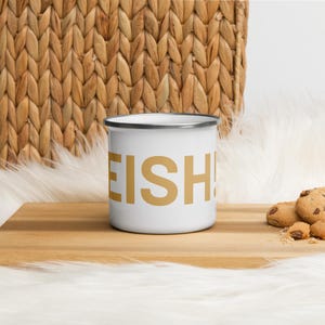 Op de afbeelding: Witte emaille camping mok met een gouden 'EISH!' afbeelding. De mok staat op een houten oppervlak.