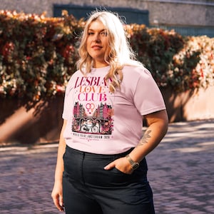 Może przedstawiać: Jasnoróżowa koszulka z napisem "LESBIAN LOVE CLUB". Grafika przedstawia most, budynki, rower i napis "WORLD PRIDE AMSTERDAM 2026". Koszulka jest noszona przez osobę.
