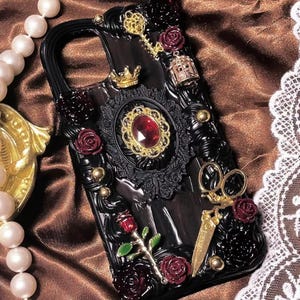 May include: Black phone case with ornate embellishments. Features a large red jewel, gold accents, and miniature roses. Includes a gold key, crown, and scissors. The case is decorated with intricate details.