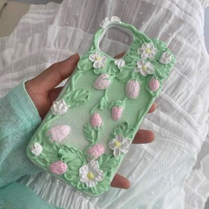 Può includere: Una custodia per telefono verde con un design 3D di fiori bianchi e fragole rosa. La custodia è realizzata per sembrare glassa.