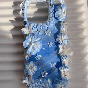 May include: A blue and white phone case with a floral design. The case is decorated with 3D flowers, stars, and butterflies. The flowers are white with a blue center and the butterflies are white with a blue outline.