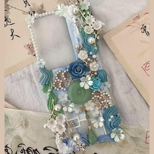 May include: A blue and white phone case decorated with pearl beads, flowers, and swirls. The case has a raised design and a clear window for the phone screen.