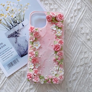 Puede incluir: Una funda de teléfono rosa decorada con flores 3D blancas y rosas. La funda tiene un aspecto texturizado y esmerilado.