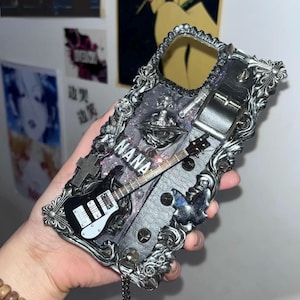 Könnte beinhalten: Eine schwarze und silberne Handyhülle mit einer silbernen Gitarre, einem silbernen Kreuz und dem Wort "NANA" darauf.