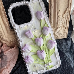 May include: A phone case featuring a 3D cream-piped border in white with small embedded pearls. The surface displays a bouquet of light purple tulips with green stems and leaves, all rendered in a textured, raised cream-piped design on a pearlescent white background.