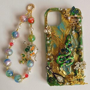 Op de afbeelding: Een sierlijke telefoonhoes en bijpassende bedel. De hoes heeft een groen en goud ontwerp met een met juwelen bezette slang, parels en bergaccenten. De bedel bevat kleurrijke kralen, parels en een decoratieve hanger met een slang en oranje accenten.