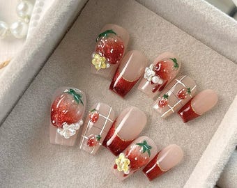 May include: A set of artificial nails with a strawberry theme. The nails feature a gradient of red glitter, with some nails showcasing 3D strawberry designs, floral accents, and a grid pattern with tiny strawberry illustrations. The nails have a glossy finish.