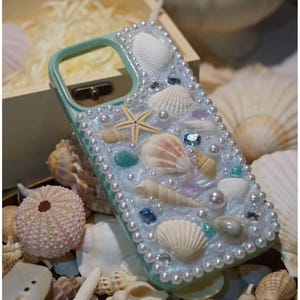 Puede incluir: Una funda de teléfono azul claro decorada con conchas marinas, perlas y otros adornos con temática playera.
