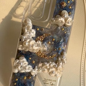 May include: A clear phone case with a night sky design. It features a blue and glittery background with a gold crescent moon, stars, and pearl-like clouds. The text "I'M ALWAYS HERE For You" is visible.
