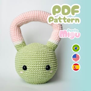 Op de afbeelding: Een gehaakt kettlebell-speeltje in lichtgroen met een roze en witte handgreep. Het speelgoed heeft zwarte ogen en kleine roze wangen. De afbeelding bevat de tekst "PDF Pattern" en de naam "Ponto Migu".