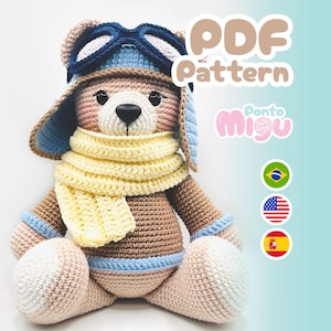 May include: A handmade crochet teddy bear wearing a pilot's hat and goggles, a yellow scarf, and blue accents. The bear is light brown, with pink and white details. The image includes the text "PDF Pattern" and the brand name "Ponto Migu".