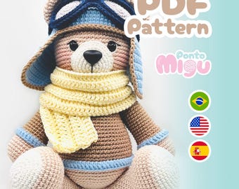 Puede incluir: Un oso de peluche tejido a ganchillo con gorro y gafas de piloto, una bufanda amarilla y detalles azules. El oso es de color marrón claro, con detalles rosas y blancos. La imagen incluye el texto "PDF Pattern" y la marca "Ponto Migu".