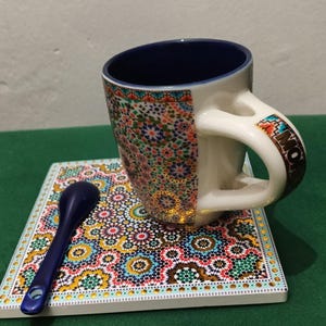 Peut inclure: Un mug en céramique avec un intérieur bleu foncé et un motif géométrique coloré à l'extérieur, portant l'inscription MOROCCO sur l'anse. Le mug est posé sur un dessous de verre carré assorti à côté d'une petite cuillère bleu foncé.