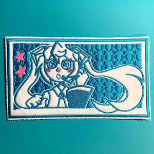 May include: Embroidered patch featuring a cartoon character with long white hair, large eyes, and pink blush. The character is set against a blue background with a star pattern. Two pink stars are in the upper left corner.