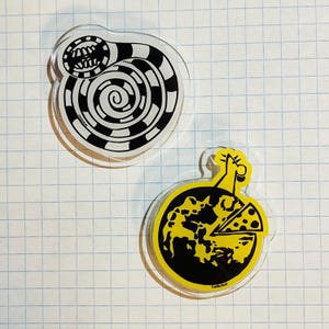 May include: Two clear acrylic pins. One pin features a black and white spiral design with a grinning mouth. The other pin has a yellow background with a black design and a slice of cheese. The pins are on a grid background.