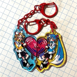 May include: Two anime-style keychains with red hardware. Each keychain features a cartoon character holding a half-heart, one in blue and yellow, the other in yellow and blue. The heart is pink with white accents.