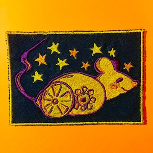 Peut inclure: Écusson brodé représentant une souris jaune avec une roue et un motif floral, sur fond bleu marine avec des étoiles jaunes et orange. Le patch a une queue et un contour violets, avec une bordure jaune.