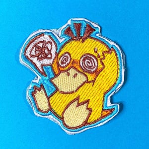 May include: An embroidered patch featuring a yellow duck-like creature with a wide beige beak, brown hair tufts, and spiral eyes. A white speech bubble containing a brown scribble is positioned to the left of the character's head.