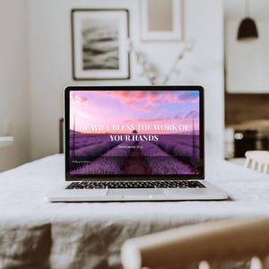 Peut inclure: Un ordinateur portable argenté affiche un écran avec le texte "HE WILL BLESS THE WORK OF YOUR HANDS" sur un champ de lavande au coucher du soleil. L'ordinateur portable est sur une nappe blanche.