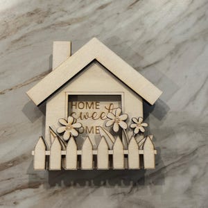 May include: A wooden house-shaped decoration with a chimney, a picket fence, and three flowers. The words "HOME sweet HOME" are visible inside the house cutout. The decoration is made of light-colored wood.