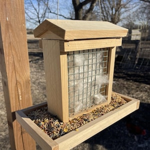May include: A wooden bird feeder with a light-colored finish, featuring a wire mesh front filled with fluffy material. The feeder has a tray filled with birdseed and a sloped roof. It is suspended outdoors.