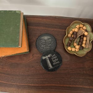 May include: Two round, dark gray slate coasters with inspirational quotes and a lighthouse design. One coaster reads "Be still, and know that I am God." The other says "Let your light shine." A stack of old books and a decorative dish with wooden beads are also present.