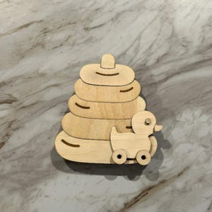 May include: A wooden stacking toy with a duck on wheels. The toy has a pyramid shape with five graduated rings. The duck has a round body, a small beak, and two wheels. The toy is made of light-colored wood.