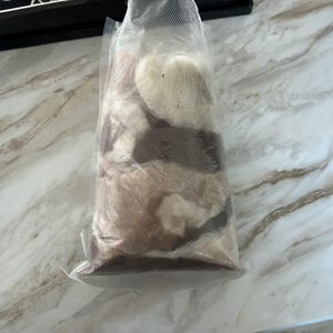 May include: A clear plastic bag filled with a mix of beige, brown, and white yarn. The yarn appears to be various textures and shades, creating a natural, earthy color palette. The bag is sealed, preserving the contents.