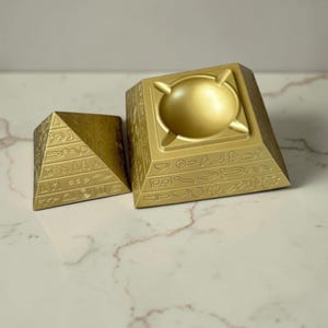 Könnte beinhalten: Zwei goldfarbene pyramidenförmige Aschenbecher. Einer ist eine kleine Pyramide, der andere eine größere Pyramide mit einem kreisförmigen Aschenbecher-Design oben. Beide haben Hieroglyphen-Schnitzereien an den Seiten.