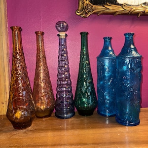 May include: Six decorative glass bottles with various colors and patterns. The bottles are arranged in a row on a wooden surface. The bottles are all different shapes and sizes.
