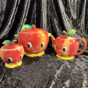 May include: A ceramic tea set featuring three apple-shaped pieces with red and yellow gradients, green leaves, and yellow bases. Each piece displays a smiling face with large eyes and eyelashes. The set includes a teapot, a lidded jar, and a bowl.