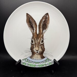 May include: Round white ceramic plate decorated with a detailed illustration of a brown hare's face. The hare has a green and blue patterned collar. The plate is displayed on a black stand, suitable for decorative or functional use.