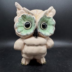 May include: A ceramic owl figurine with a light pink body, green eye accents, and black eyes and beak. The owl has a whimsical design with detailed feather-like patterns and a slightly rounded shape, standing upright.