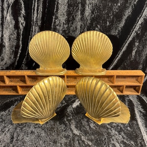May include: Four brass seashell bookends. Each bookend is a detailed, fan-shaped scallop shell design with a flat base. The bookends are a warm gold color and sit on a dark background.