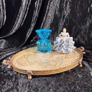 May include: An oval, gold-toned metal tray with ornate floral detailing and decorative handles. A blue, geometric perfume bottle and a small figurine in a ruffled dress are displayed on the tray, set against a dark, textured backdrop.