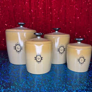 May include: A set of four vintage kitchen canisters in a cream and tan ombre design. Each canister has a black label with a decorative border and the words "Flour", "Coffee", "Sugar", and "Tea". The canisters have black knobs on the lids.