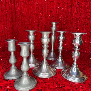 May include: A collection of six silver-toned candlesticks with varying designs. The candlesticks have a classic, elegant appearance, with round bases and detailed stems. They are arranged on a red sequined surface.