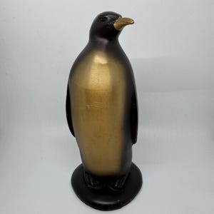 May include: A standing penguin figurine featuring a smooth, metallic gold finish on the chest and a dark brown, almost black, finish on the head, back, and base. The penguin has a pointed, light gold beak and is positioned on a circular base.